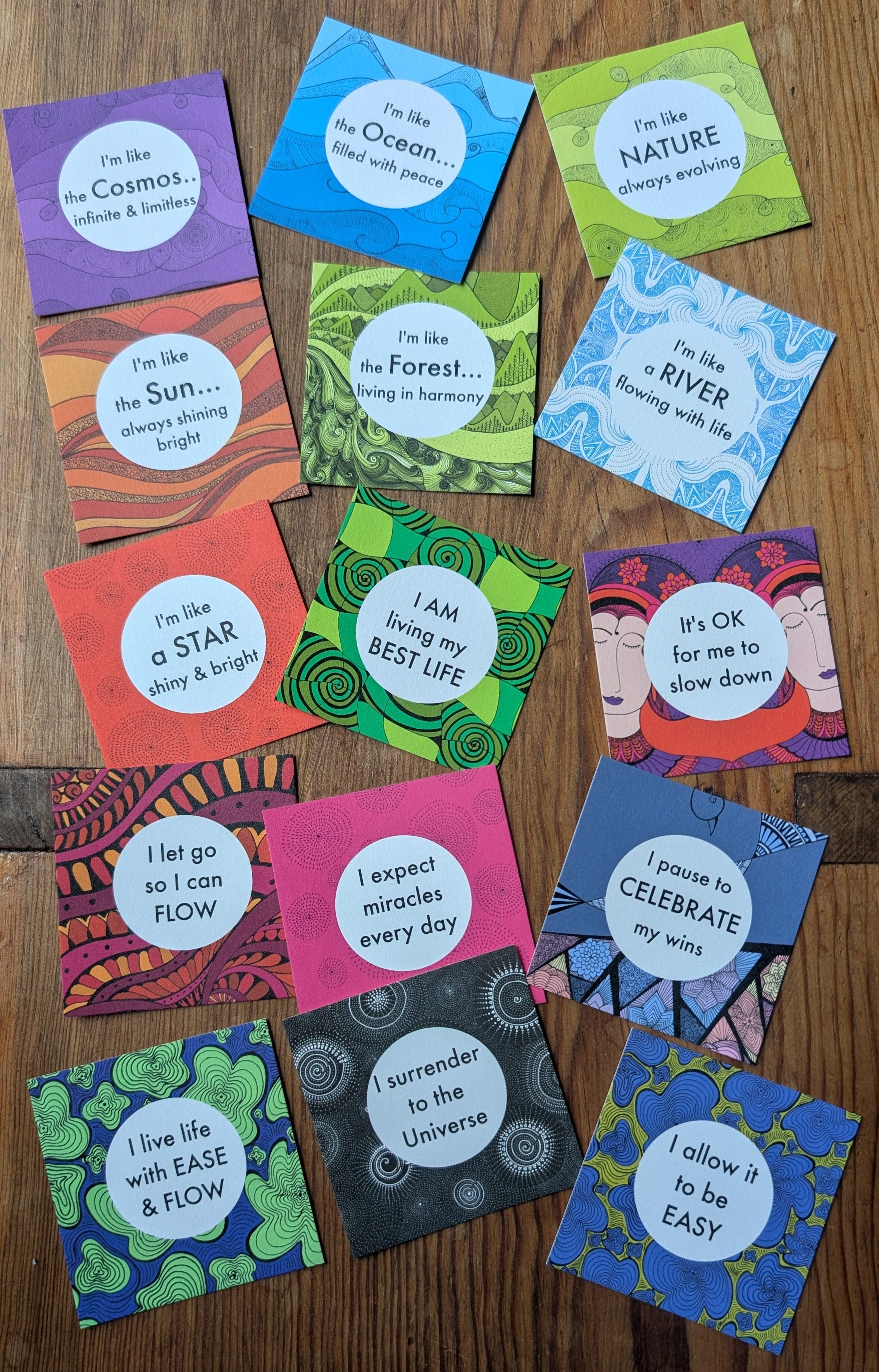 A set of colorful square printed cards or booklets arranged in a circle on a wooden tabletop. Each card has a white oval label with handwritten-style text, and the cards feature bright multicolor abstract and illustrated designs.