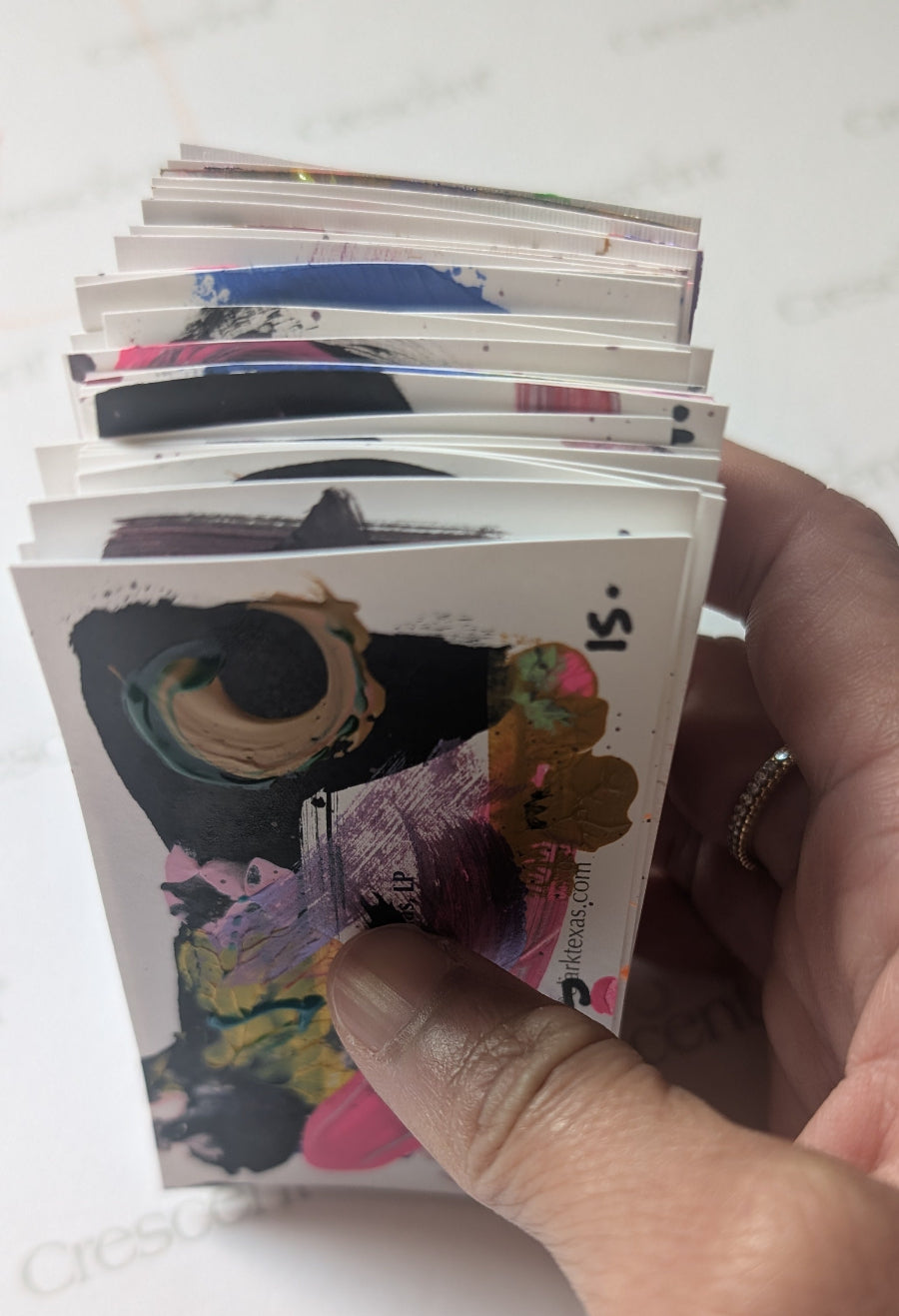 Hand holding a stack of colorful greeting cards with a visible brand name on a white surface.