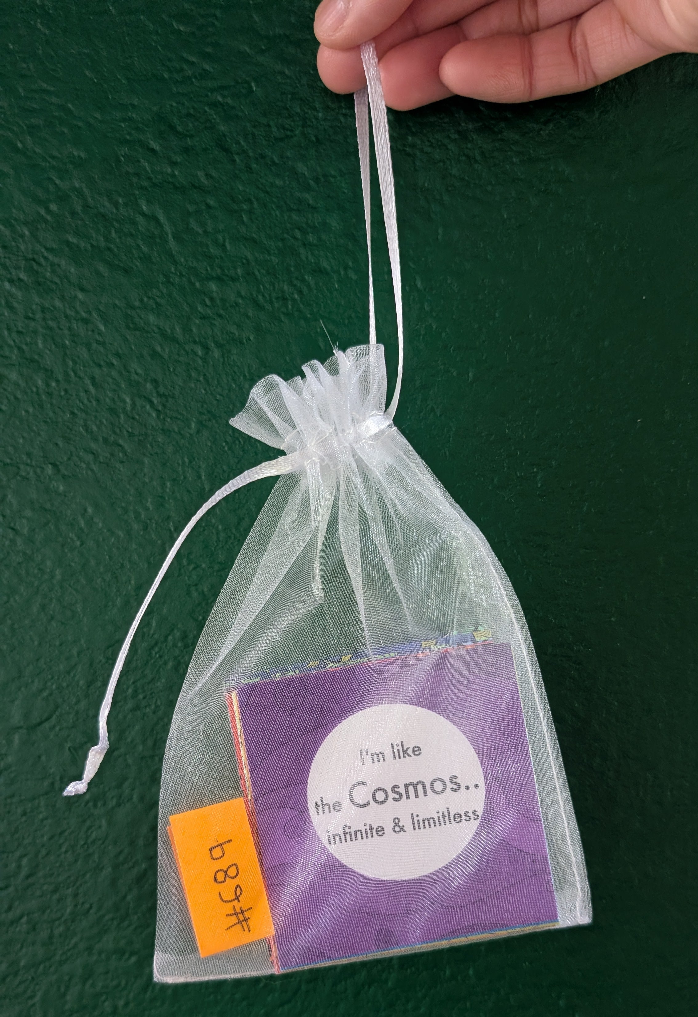 A hand holds a clear organza drawstring pouch containing a purple square card that reads 'I'm like the Cosmos.
