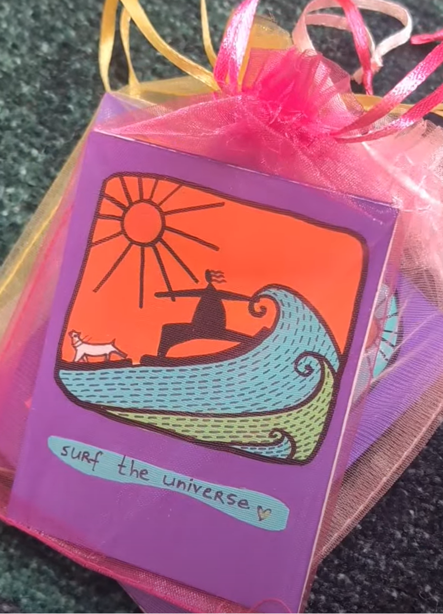 Card with a design of a person surfing a wave, sun, and dog, wrapped in pink tissue paper.